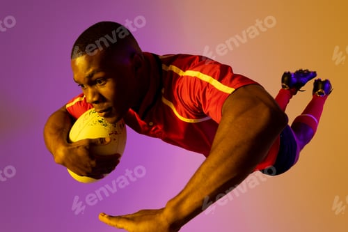 Preview: African american male rugby player jumping with rugby ball over pink lighting