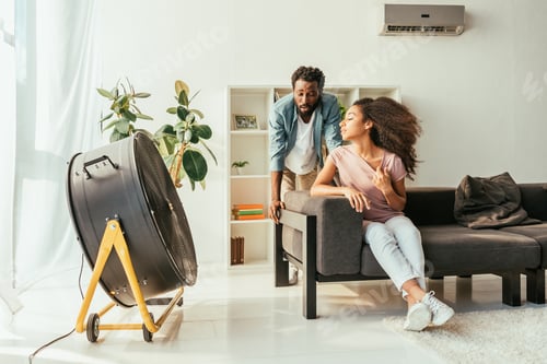 Preview: exhausted african american man and woman suffering from summer in front of blowing electric fan