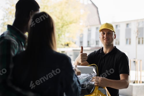 Preview: Delivery man in yellow uniform delivering parcel box to recipient - young woman and her husband