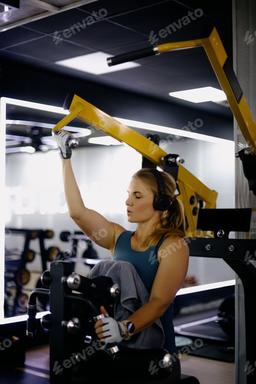 Preview: Young caucasian female exercising with machine in gym wearing headphones
