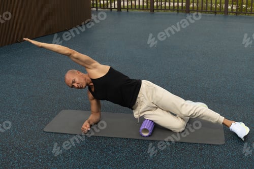Preview: Athlete man stretching legs on foam roller on sports ground. Outdoor. Morning routine.