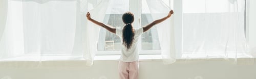 Preview: back view of african american girl in pajamas opening window curtains in sunshine, horizontal image