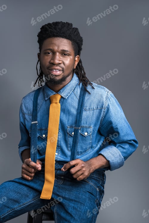 Preview: handsome african american man sitting on chair and holding suspenders isolated on gray