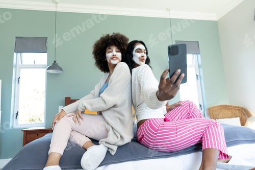 Preview: Biracial friends with cream on faces posing and taking selfie over smartphone while sitting on bed