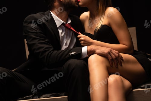 Preview: cropped view of sexy woman holding tie of man in bed in dark room