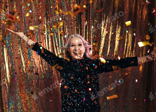 Preview: Smiling senior woman in black dress having fun under colorful confetti against a golden backdrop