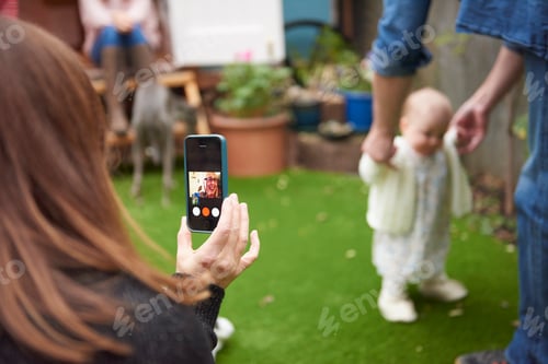 Preview: Mother in garden using smartphone to film daughter learning to walk, focus on foreground