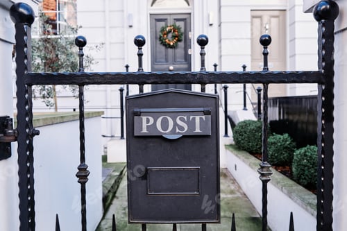 Preview: Closeup of an aged rusty black post box near a house with a blurry background