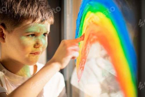 Preview: Smiling cute european small kid draws rainbow on window with finger and paint, close up, sun flare