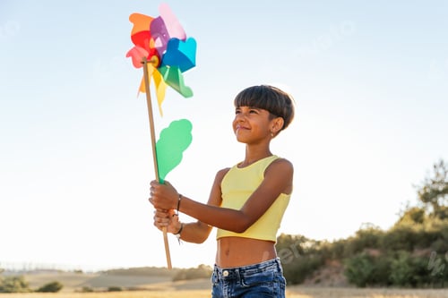 Preview: Young girl holding a colorful wind turbine toy standing outdoors in a field.