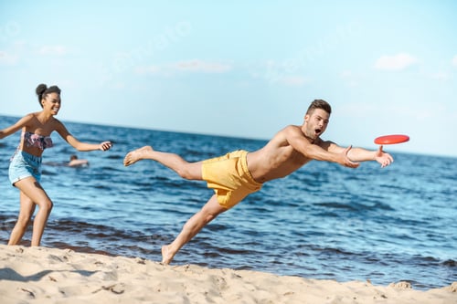 Preview: young multicultural couple playing with flying disc on beach on summer day
