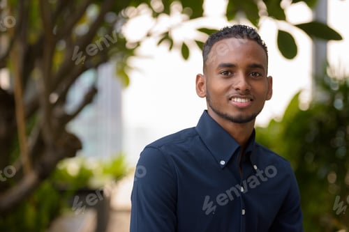 Preview: Handsome African businessman sitting outdoors at rooftop garden and smiling