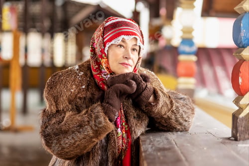 Preview: A Slavic woman in a national scarf,a fur coat and warm mittens on the porch of a wooden house