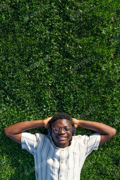 Preview: Top view of black man relaxing, lying on grass listen to music with headphones. Copy space. Vertical