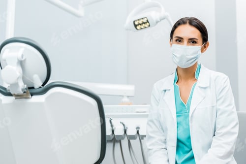 Preview: female dentist in white coat and mask near dental chair