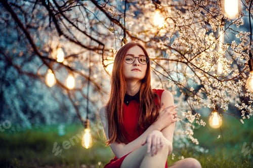 Preview: Ginger long haired young girl in glasses sits on grass under blossom white tree, evening light lamps