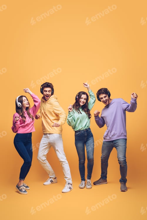 Preview: positive friends listening music in headphones and dancing together, on yellow