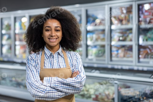 Preview: Smiling supermarket employee in apron stands with arms crossed in front of refrigerated section