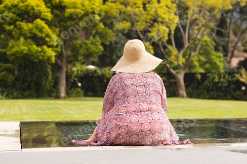 Preview: Senior biracial woman sits by a pool, wearing a wide-brimmed hat and a floral dress