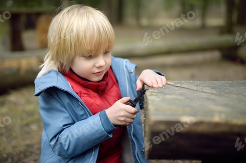 Preview: Little boy saws log with a small penknife during walking in the forest at spring or autumn. Outdoor