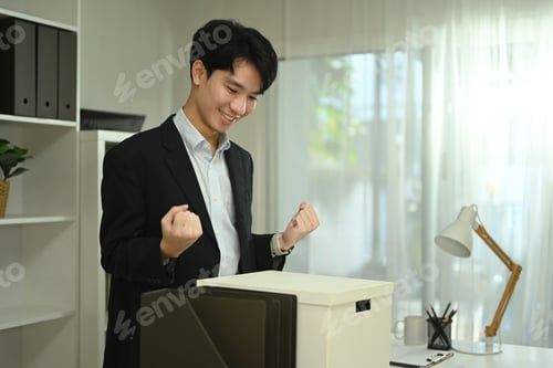 Preview: Happy asian male office worker with box of his belongings moving into a new office