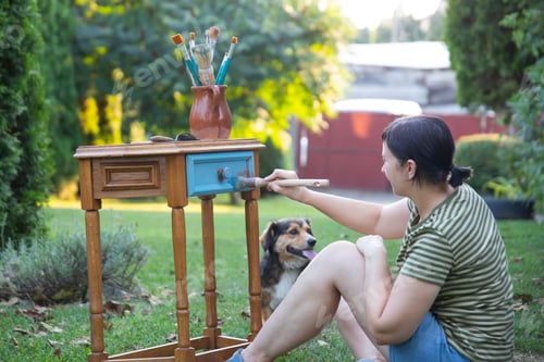 Preview: Woman painting a wooden furniture while sitting with her dog in the garden. DIY concept.
