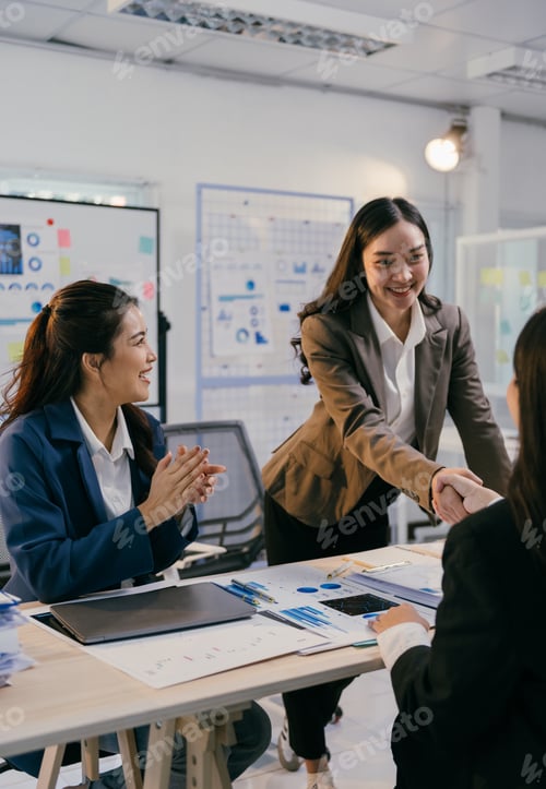 Preview: Businesswomen shaking hands celebrating successful agreement in modern office with charts and graphs