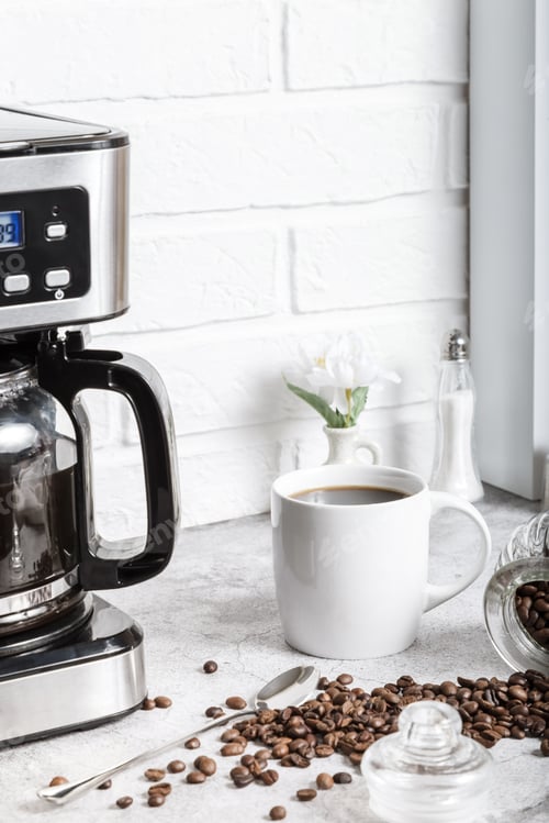 Preview: A white cup with a drink on the kitchen table next to a drip coffee maker. Roasted coffee beans.