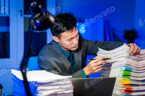 Preview: A young man works at his desk late at night in the office, surrounded by many documents. Checking