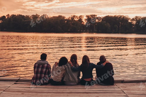 Preview: Among best friends. Rear view of young people in casual wear embracing while sitting on the pier