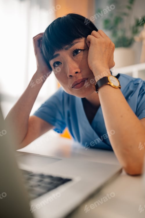 Preview: Asian female medical professional seated at desk with hand on forehead