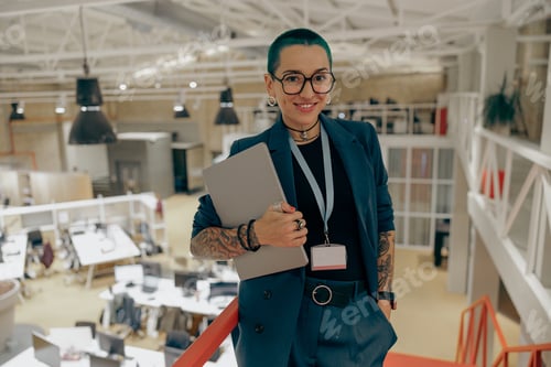 Preview: Smiling tattooed business woman with green short hair holding laptop standing in modern office