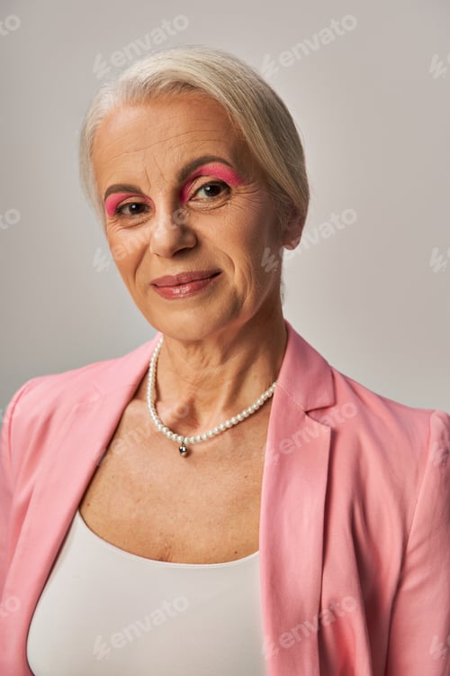 Preview: portrait of elegant senior lady in pink blazer and pearl necklace smiling at camera on grey