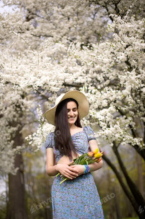 Preview: A young girl in a hat and with tulips, standing in a blue dress next to a blooming white tree
