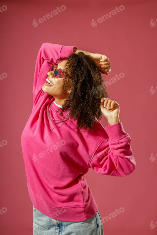 Preview: Woman in heart-shaped glasses with raised hands looking at side standing on pink studio