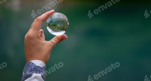 Preview: A woman's hand holds a spherical glass ball against the background of a beautiful mountain lake.