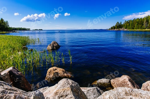 Preview: Clear water blue lake with stones and green forest on a sunny summer day in Finland.