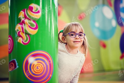 Preview: Cute little girl in eyeglasses standing behind the column with painted lollipops and candies.