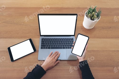 Preview: Hands holding a blank white screen mobile phone and laptop with black tablet on wooden table