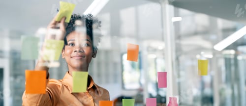 Preview: Happy black businesswoman planning new porject with sticky note in a creative office