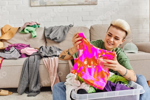 Preview: young woman with cheerful smile sorting thrift store finds, holding colorful top near plastic
