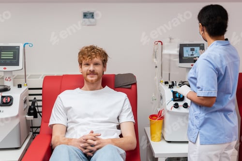 Preview: multiracial nurse in blue uniform operating transfusion machine near plastic cup and redhead