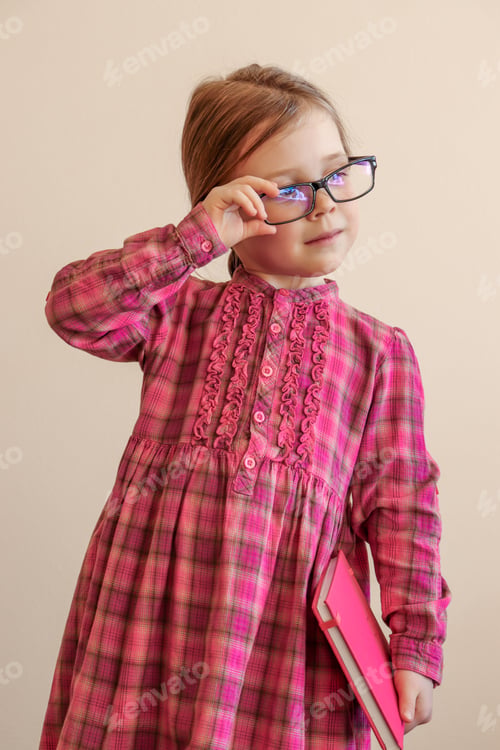 Preview: A 6-year-old girl in a pink dress and glasses stands and holds a sketchbook in her hands