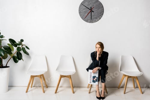 Preview: bored young businesswoman holding newspaper and sitting with hand on chin in queue