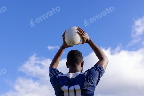 Preview: African American young male athlete holding white rugby ball above head on field, copy space