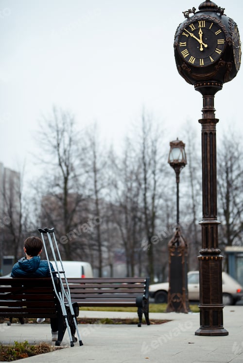 Preview: A physically disabled young man is sitting on a bench near a street clock, crutches nearby.