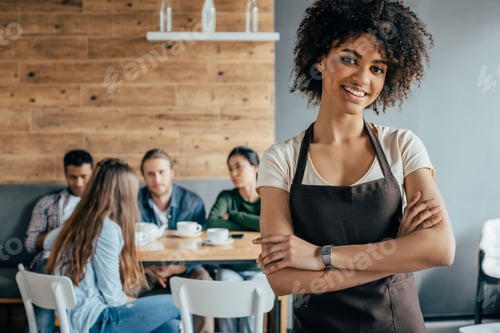 Preview: Smiling african american waitress standing with customers sitting behind in cafe
