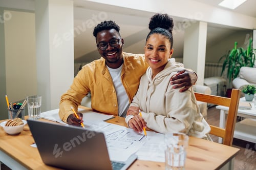 Preview: Diverse couple using laptop and looking into the blueprints of their new home