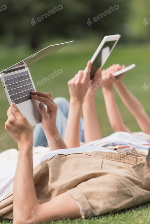 Preview: cropped shot of family using digital devices while lying on lawn in park