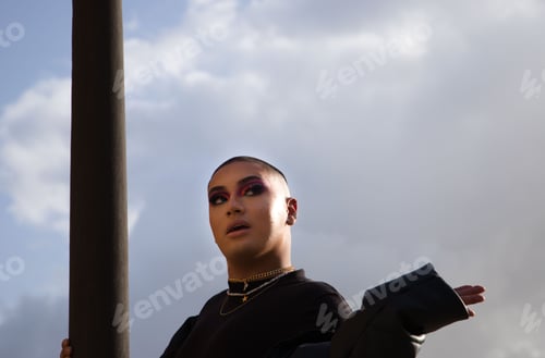 Preview: Young attractive Latin non-binary person wearing makeup posing outdoors against a cloudy sky